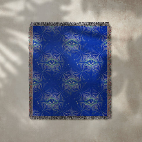 Woven cotton throw blanket with a deep blue background featuring a repeated pattern of all-seeing eyes in gold and seafoam green. The blanket has a colorful fringed edge.